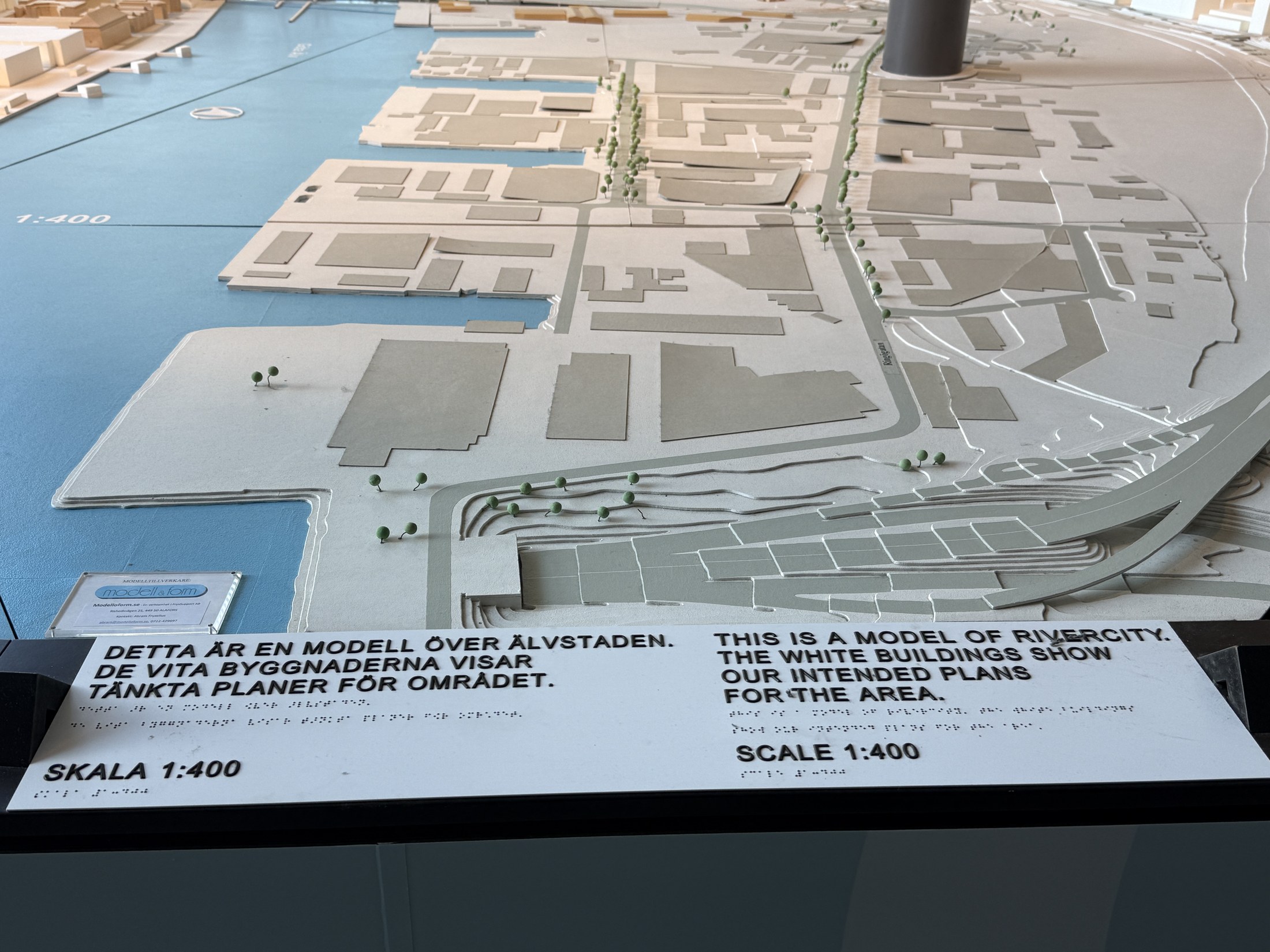 The caption board of the Älvstaden city model in Gothenburg, in Swedish, English and braille: This is a model of RiverCity. The white buildings show our intended plans for the area. Scale 1:400. Behind it, the model stretches out with the river in light blue.